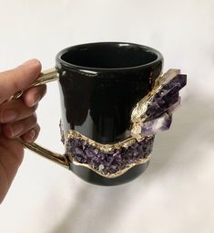 a hand holding a black coffee mug with gold trimmings and amethyst crystals