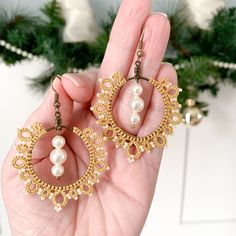 a hand holding two pairs of gold and pearl earrings in front of a christmas tree