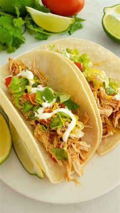 two shredded chicken tacos on a plate with limes, tomatoes and cilantro