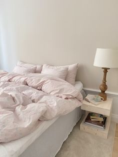 an unmade bed with pink sheets and pillows in a white room next to a lamp