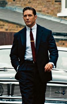 a man in a suit and tie walking next to a car with his hands in his pockets