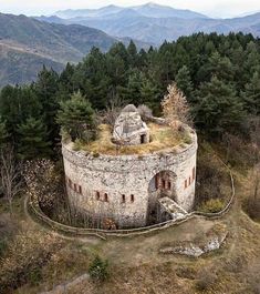 an old stone tower with grass on top in the middle of trees and mountains behind it