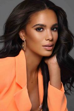 a woman with long hair wearing an orange top and gold hoop earrings, posing for the camera