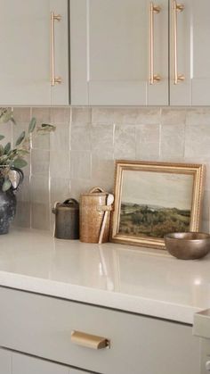 a kitchen counter with white cabinets and gold pulls on the handles, along with a framed painting