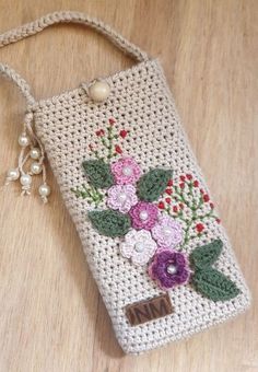 a crocheted cell phone case with flowers and pearls hanging from the side on a wooden surface