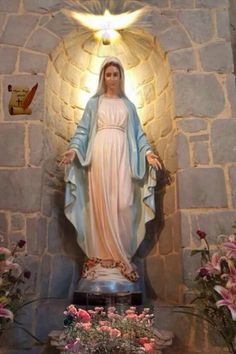 a statue of the virgin mary in front of a stone wall with flowers around it