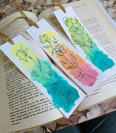 three bookmarks with flowers painted on them sitting on top of an open book,