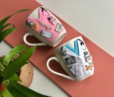 two coffee mugs with different designs on them sitting next to each other near a potted plant