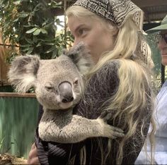 a woman holding a koala in her arms while standing next to a man with long blonde hair