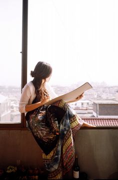 a woman sitting on top of a window sill holding a book and looking out at the city