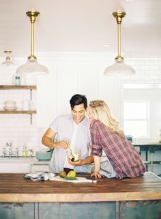 a man and woman sitting at a kitchen counter cutting vegetables together, with lights hanging above them