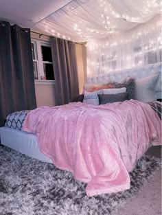 a bed covered in pink and grey blankets next to a window with lights on it