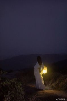 a woman in white dress holding a lantern on top of a hill at night with mountains in the background