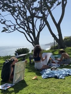 two women sitting on the grass painting in front of some trees by the water with their backs to the camera