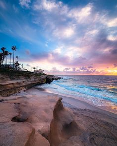 the sun is setting over the ocean with palm trees on the shore and rocks in the foreground