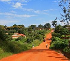 two people walking down the middle of a dirt road with houses on either side and trees in the background