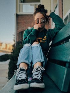 a woman sitting on top of a green bench next to a tall brick building and wearing glasses