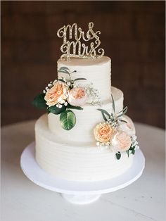 a wedding cake with flowers and mr and mrs topper on the bottom tier is sitting on a white table