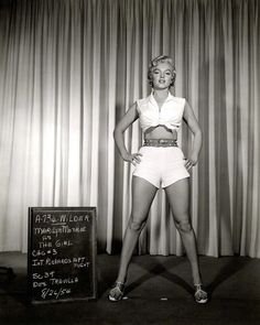 an old photo of a woman in short shorts standing next to a chalkboard with writing on it