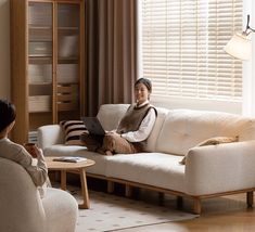 a woman sitting on a couch in front of a window using a laptop computer while another person looks at her