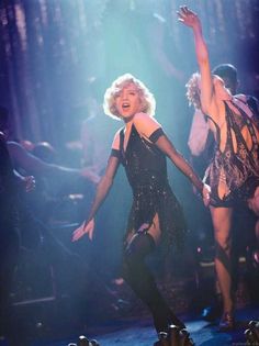 the blond haired woman is dancing on stage with other people in the background and lights behind her