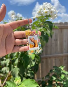 a person holding up a small photo keychain in front of a fence and flowers