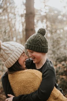 two people are hugging each other in the woods with trees behind them and one person is wearing a beanie