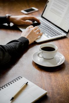 a person typing on a laptop computer next to a cup of coffee and notebooks