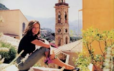 a beautiful woman sitting on top of a roof next to a tall tower with a clock