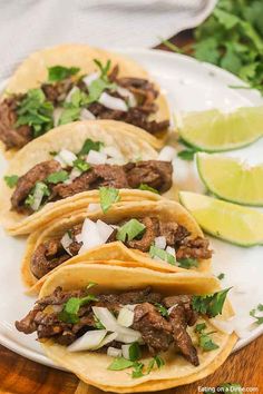 three tacos on a plate with limes and cilantro in the background