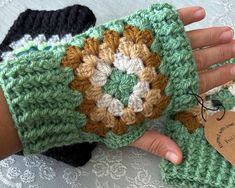 a hand holding a crocheted green and white wrist warmer with a tag on it