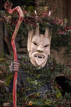 a person with a mask on holding a stick in front of some bushes and plants