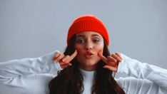 a young woman wearing an orange beanie making a funny face with her hands and fingers