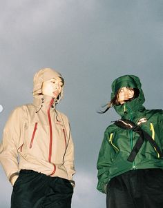 two people standing next to each other in front of a cloudy sky and one is wearing a green jacket