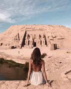 a woman in a white dress sitting on the edge of a cliff looking at an ancient building