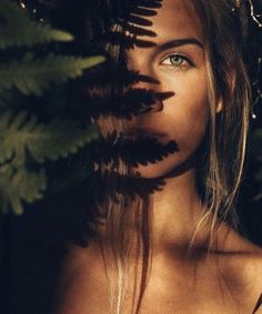 a woman with long hair and blue eyes is standing in front of some leaves, looking at the camera