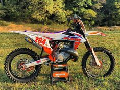 an orange and white dirt bike parked on top of a grass covered field with trees in the background