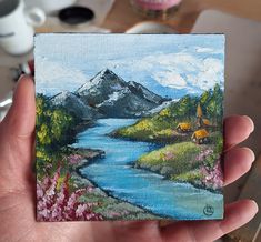 a hand holding up a small painting of a mountain lake and flowers in the foreground