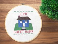 a cross stitch house with the words home sweet home on it next to a potted plant