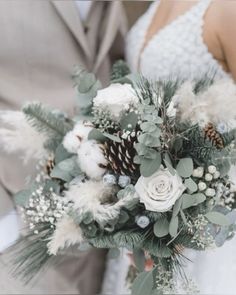 a bride and groom holding a wedding bouquet with white flowers, greenery and pine cones