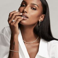 a woman with long black hair wearing a white shirt and pearl necklace, holding her hand to her mouth