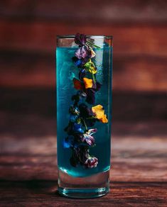 a tall glass filled with blue liquid and colorful flowers on the rim, sitting on a wooden table