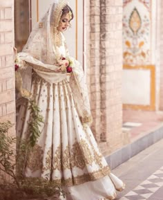 a woman in a white and gold bridal gown is standing by a brick wall