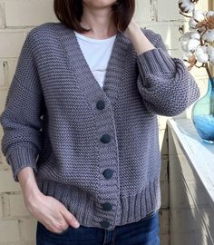 a woman standing in front of a white brick wall wearing a gray cardigan sweater