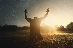 a man is standing in the grass with his hands up to the stars above him