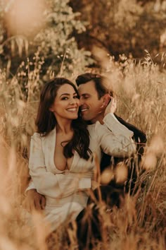 a man and woman are sitting in tall grass smiling at each other as they pose for the camera