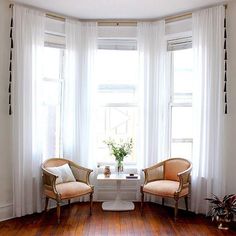 two chairs and a table in a room with white curtains on the windowsills