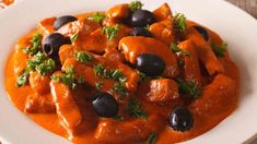 a white plate topped with chicken and olives covered in sauce on top of a wooden table