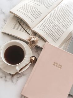 an open book next to a cup of coffee on a marble table with a spoon