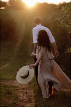 a man and woman walking down a dirt road holding hands with the sun setting behind them